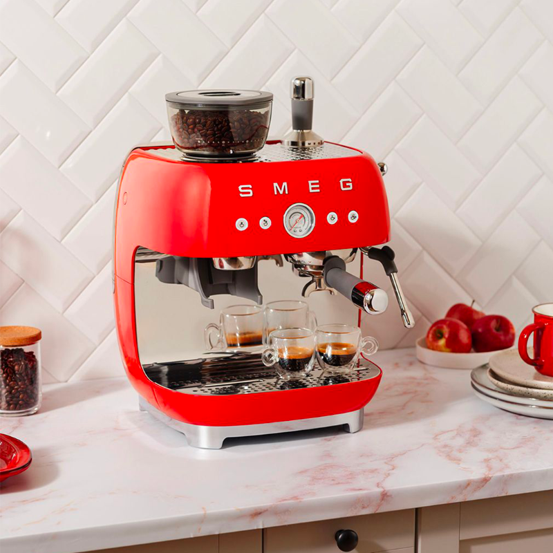 Red SMEG espresso machine on a kitchen counter with coffee cups and apples.